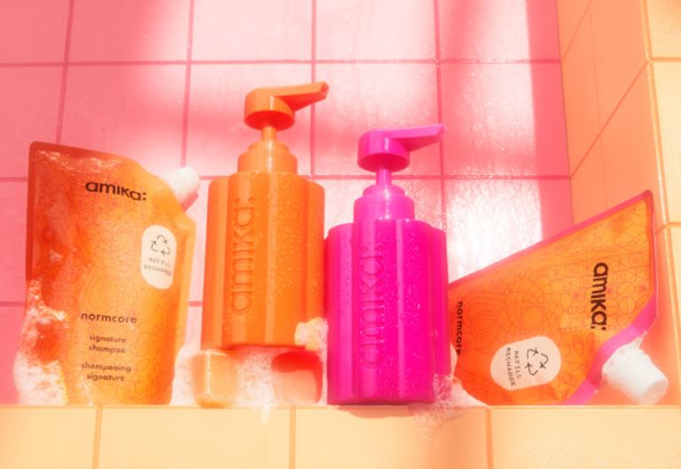 Orange and pink amika reusable pump bottles displayed with flexible haircare refill pouches in a tiled bathroom setting.