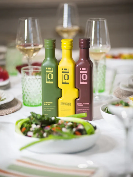 Three Foli condiment bottles in green, yellow and burgundy displayed beside a salad on a dining table.