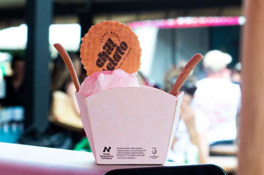 Pink paper ice cream pot containing ice cream with two spoons, displayed at an event.