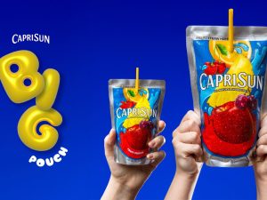 Hands holding standard and oversized Capri Sun fruit drink pouches against a blue background.
