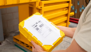 Person holding a rectangular parcel package with a yellow reinforced outer frame, white central panel and shipping label, beside yellow industrial equipment.